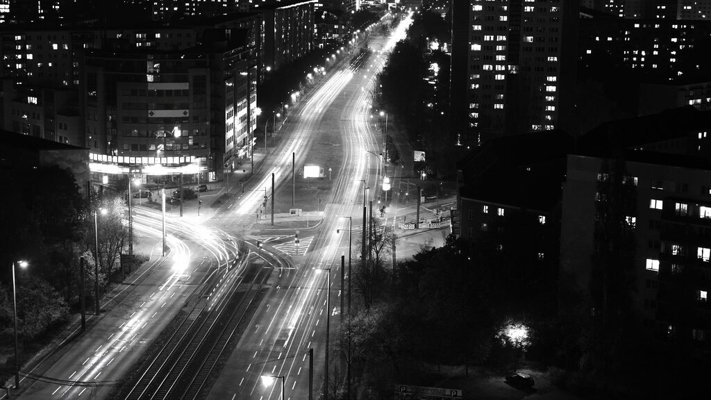 Una foto in bianco e nero di una strada cittadina di notte, con le luci dei veicoli che creano scie luminose su un incrocio, circondato da edifici residenziali illuminati.